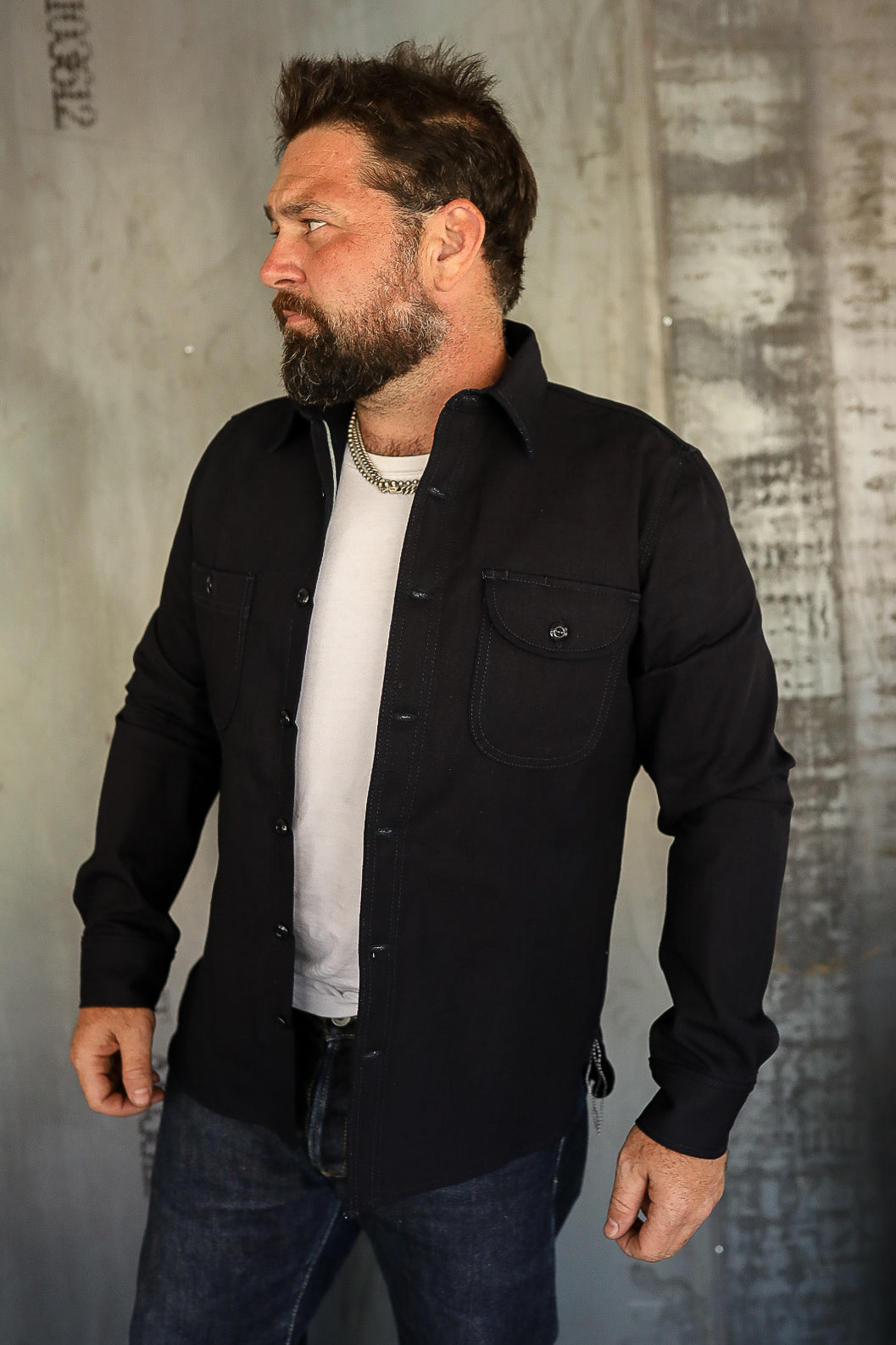 Bearded man wearing black button-up shirt over white tee and dark jeans in industrial setting