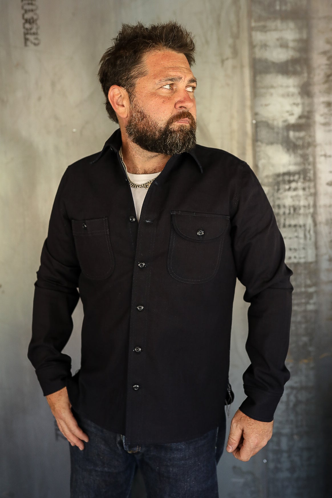 Bearded man wearing black button-up shirt with chest pockets and dark jeans, standing against industrial wall