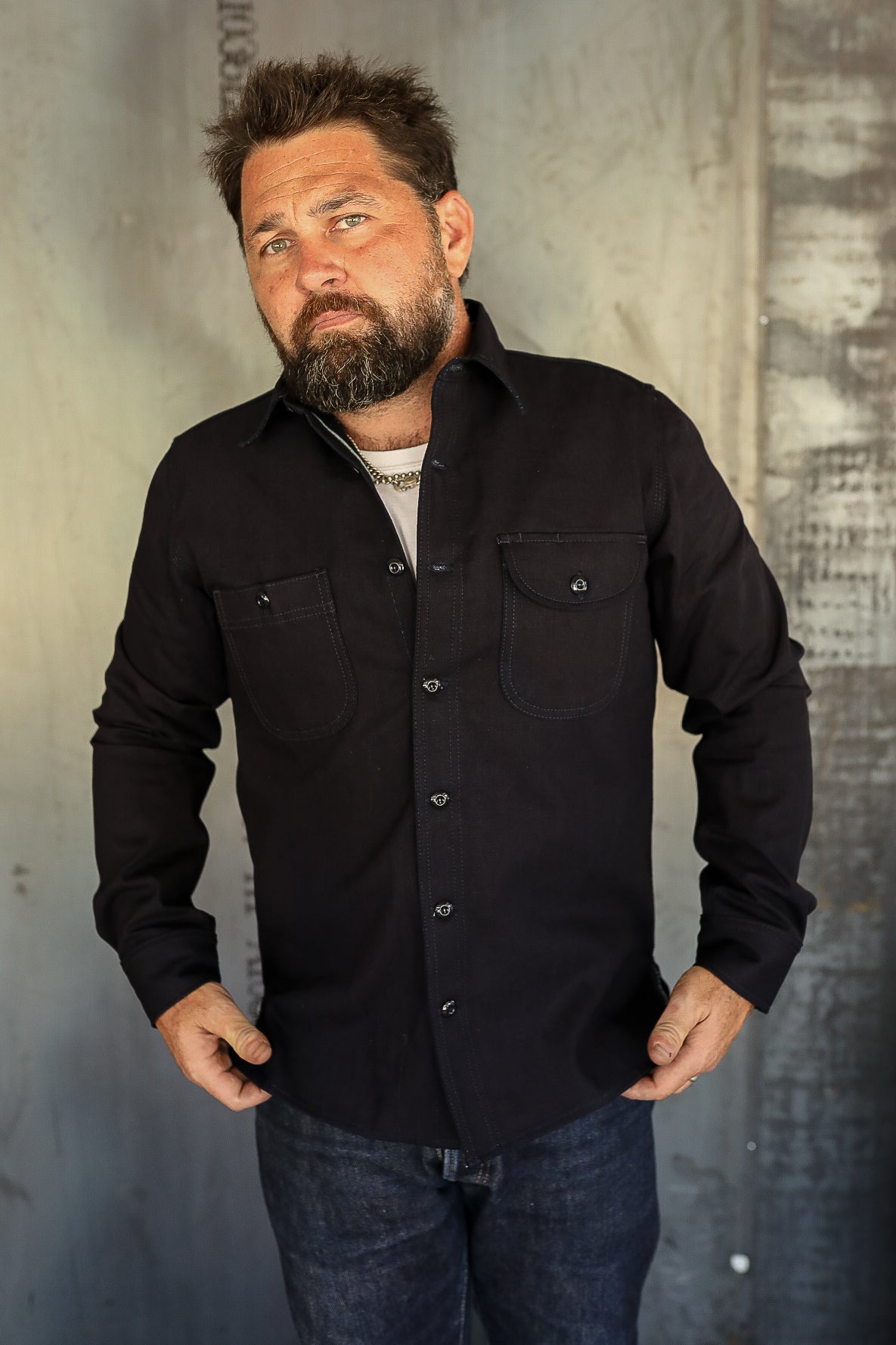 Man wearing black button-up work shirt with chest pockets standing against textured industrial wall