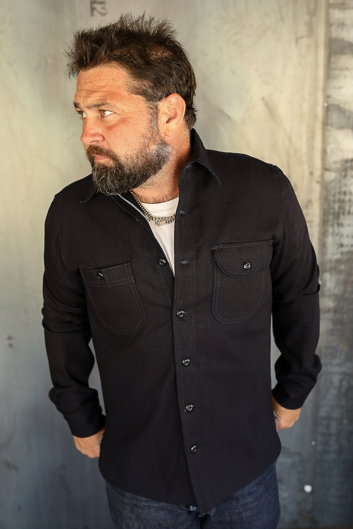Bearded man wearing a black button-up work shirt with chest pockets, industrial background