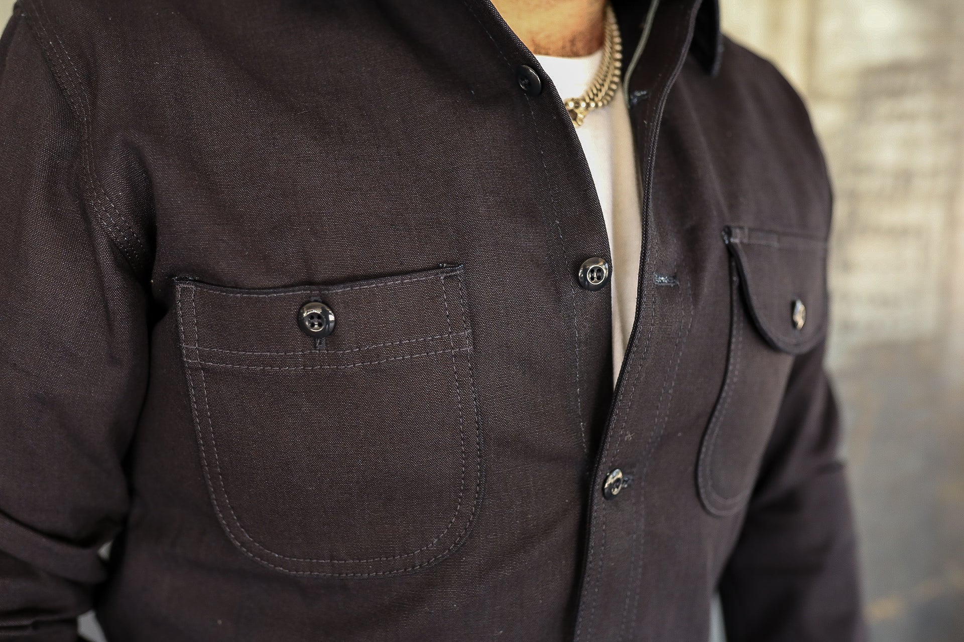 Close-up of a dark brown work shirt with buttoned chest pockets and rolled sleeves, worn over a white shirt with a gold chain necklace
