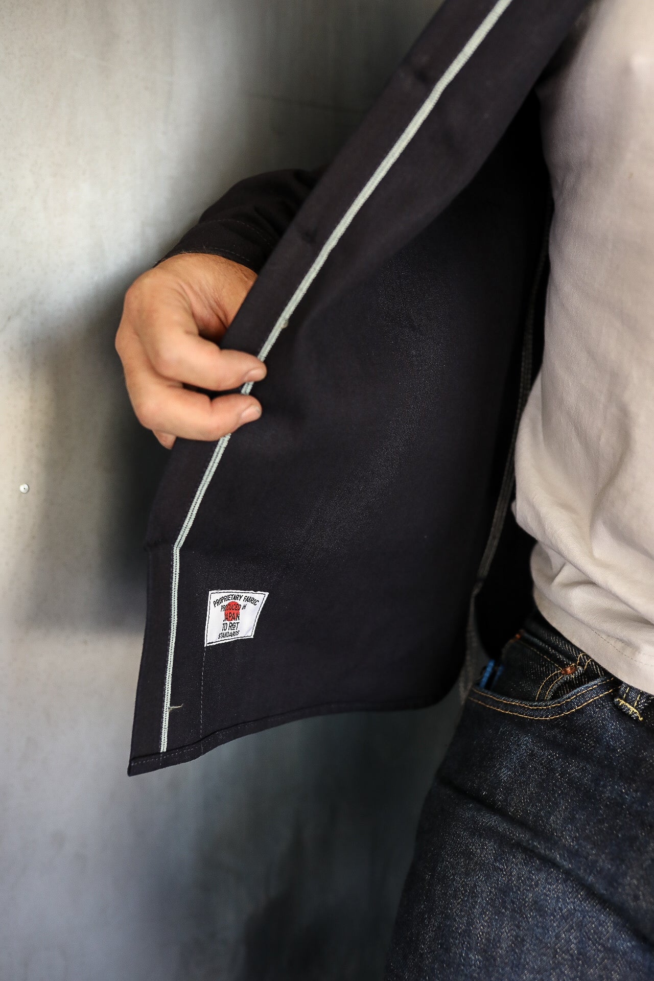 Close-up of person wearing dark jacket with white piping and tag reading 'Produced in Japan' over white shirt and jeans
