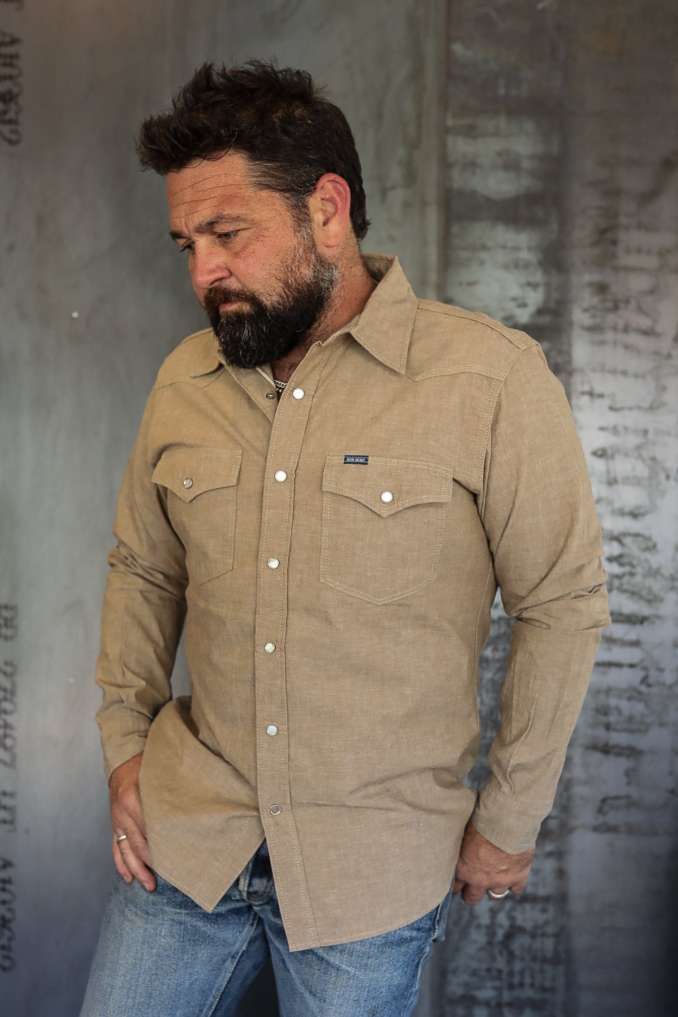 Man wearing tan button-up work shirt with chest pockets and blue jeans in industrial setting
