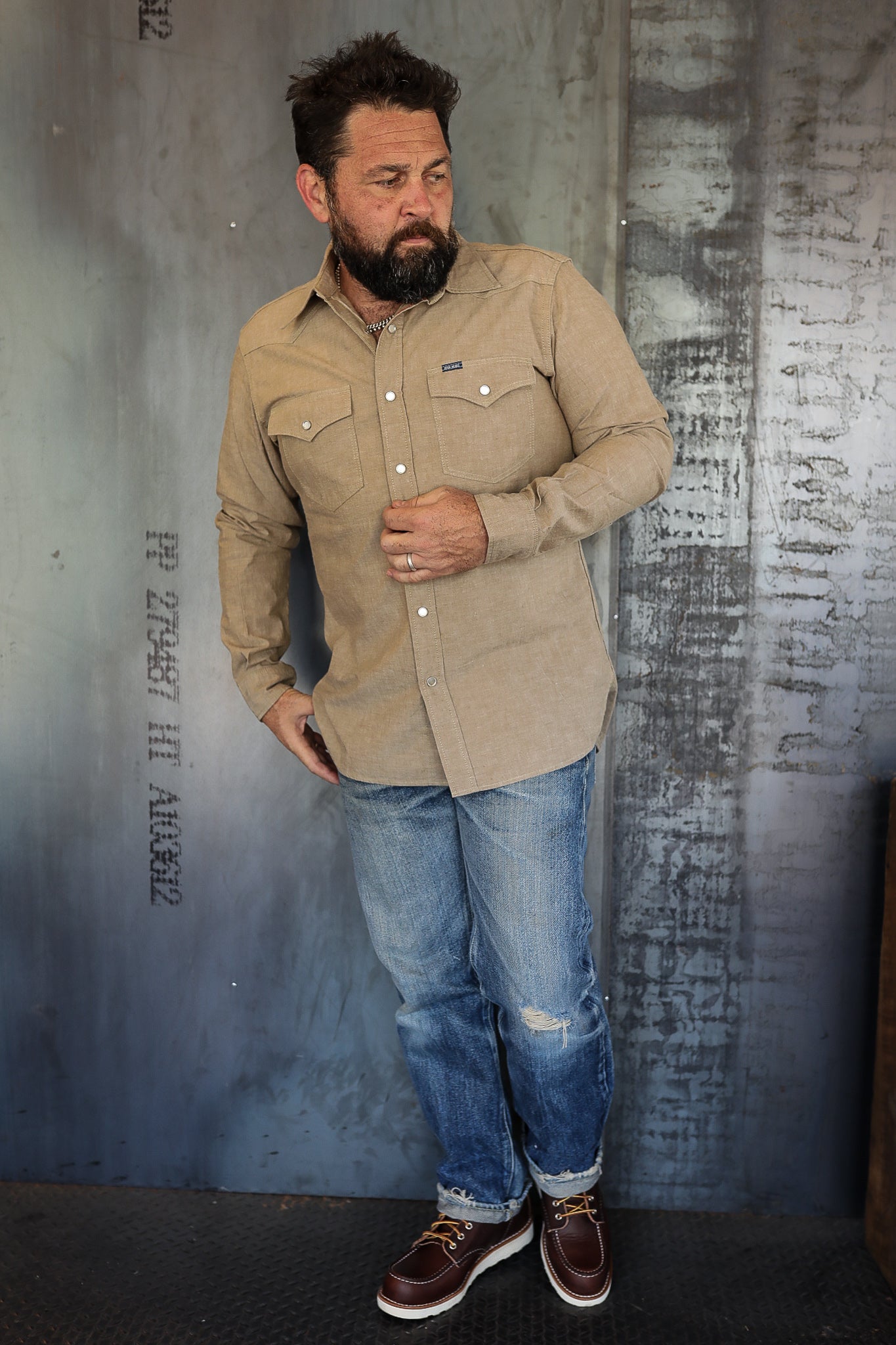 Bearded man in tan button-up shirt, distressed blue jeans, and brown leather boots standing against industrial metal wall