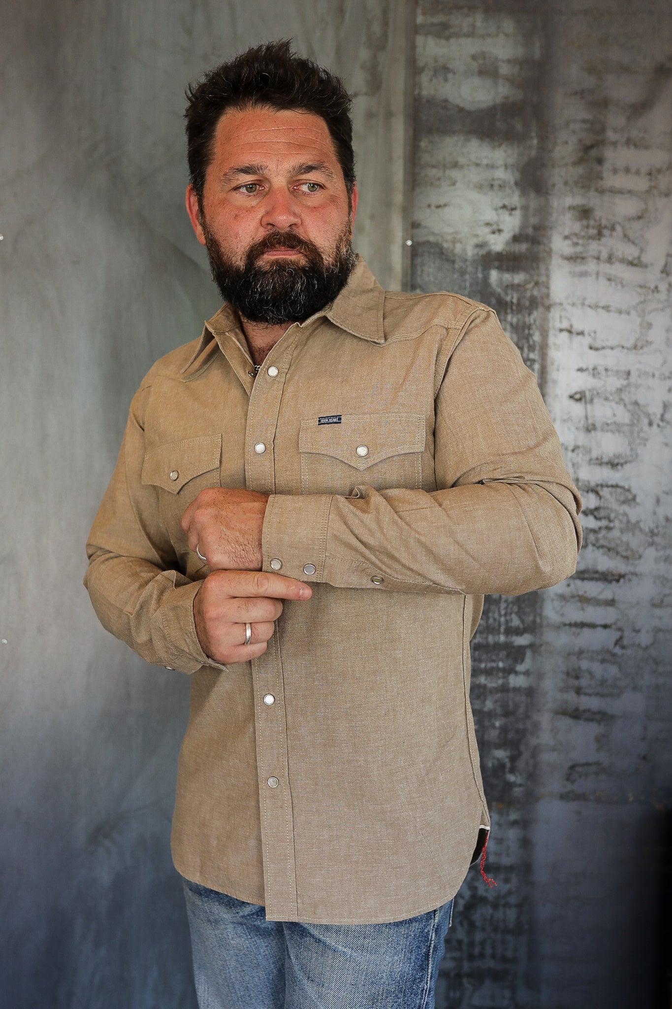 Bearded man wearing tan button-up work shirt from Iron Shop Provisions adjusting sleeve in industrial setting
