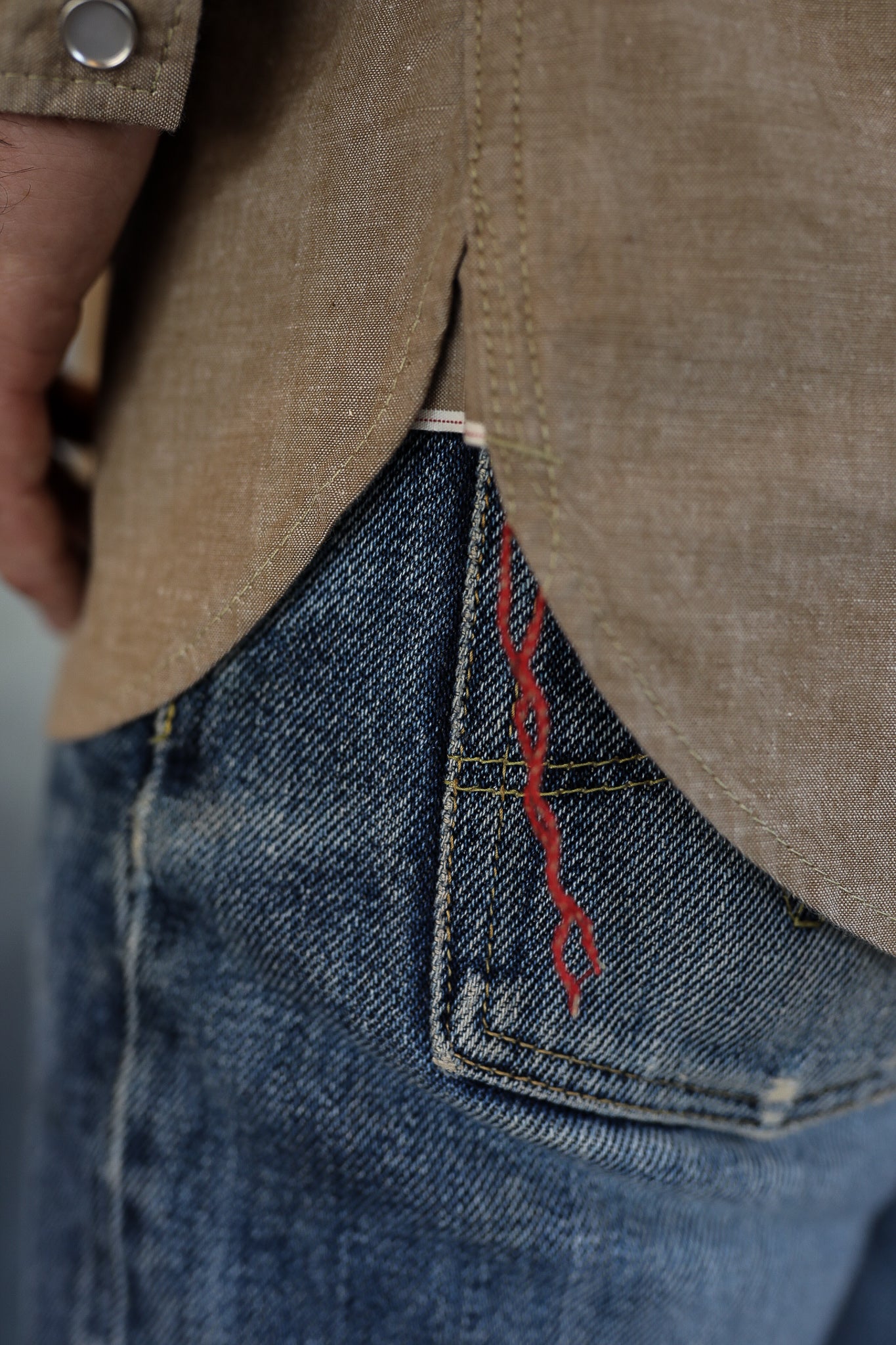 Close-up of blue denim jeans pocket with red thread detail under a brown shirt hem with metal snap button