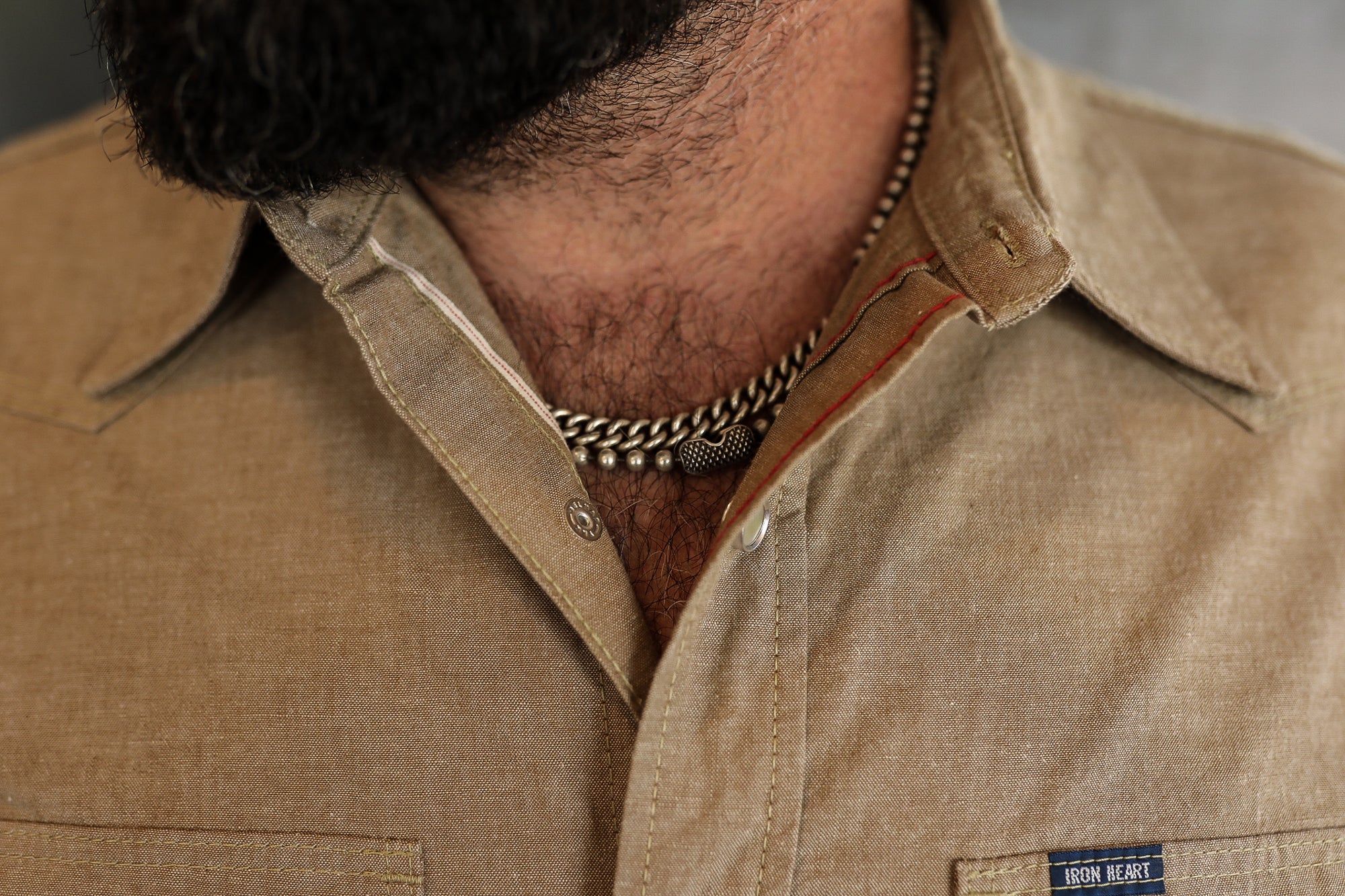 Close-up of a man wearing a tan Iron Heart shirt with silver chain necklaces and visible chest hair