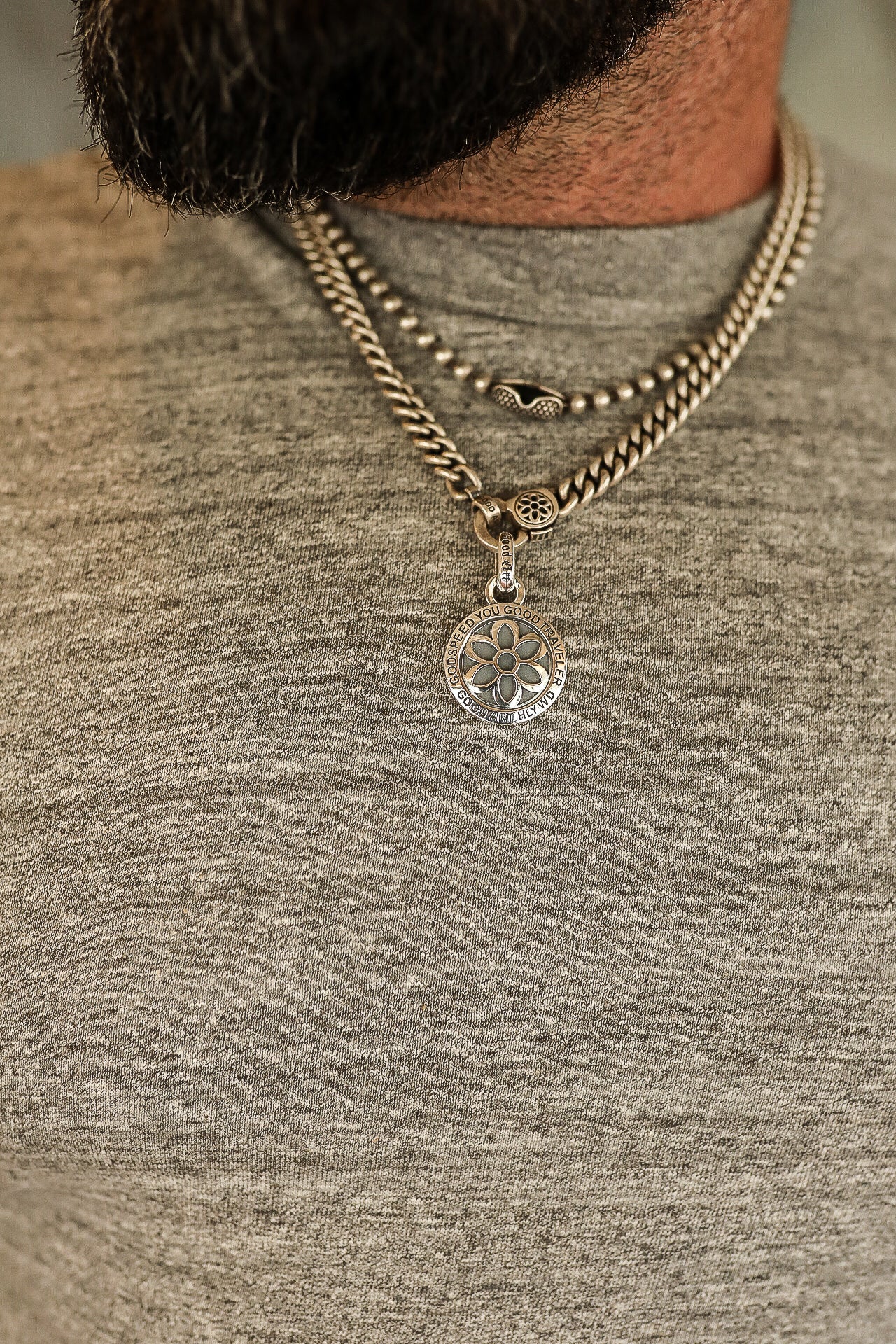 Close-up of a man wearing layered silver chains with a floral pendant and engraved round charm on a gray shirt