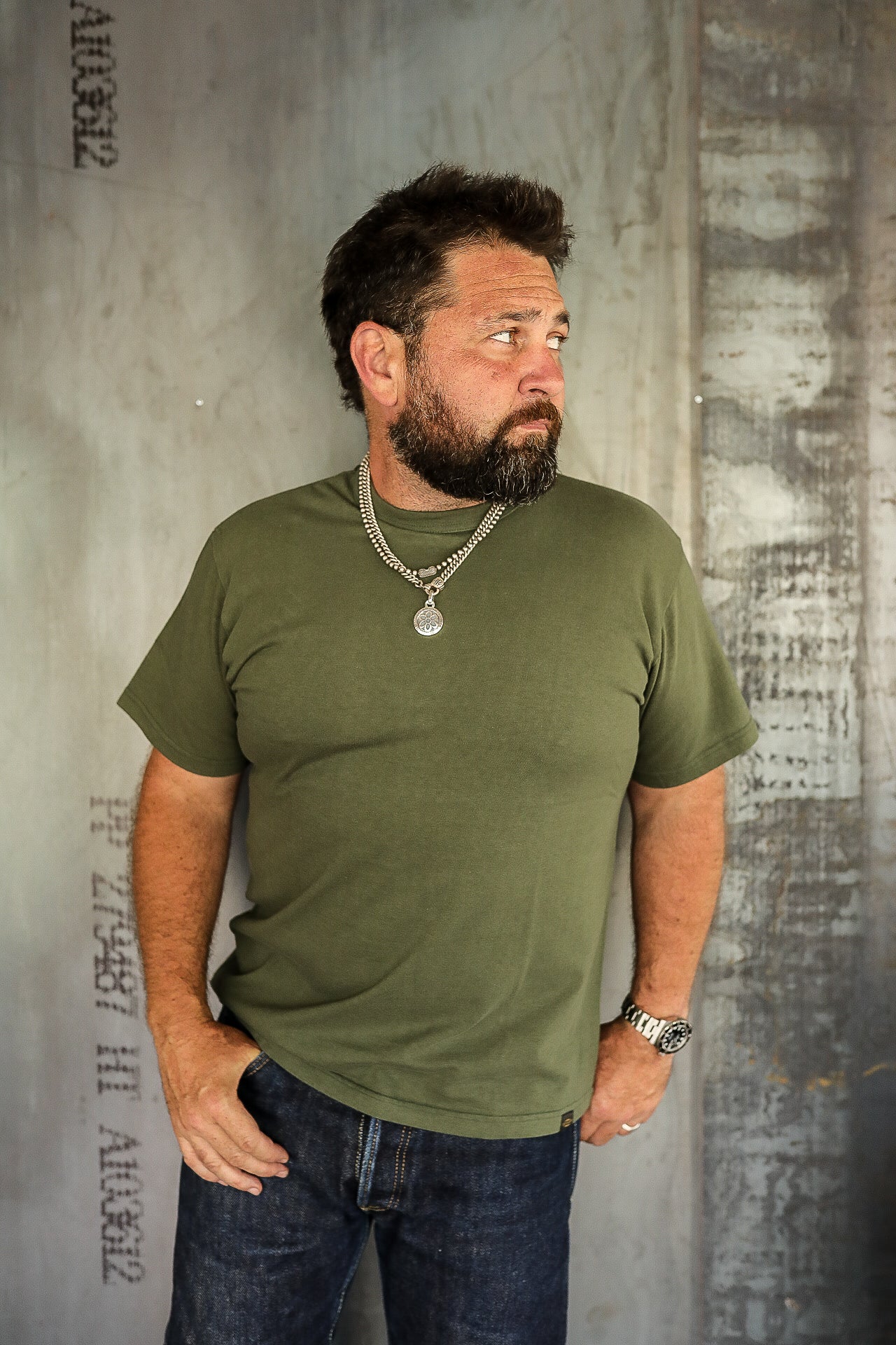 Bearded man wearing olive green t-shirt and dark jeans standing against industrial metal wall