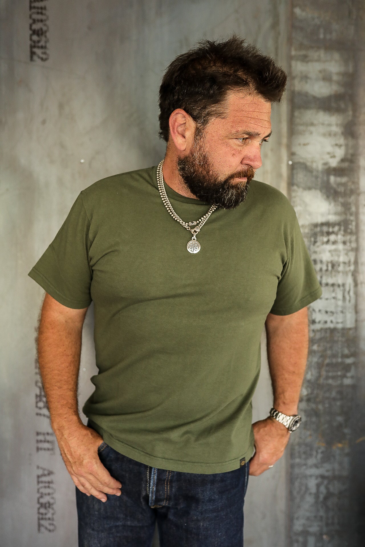 Bearded man wearing olive green t-shirt, blue jeans, silver necklace with pendant, and wristwatch against rustic metal background