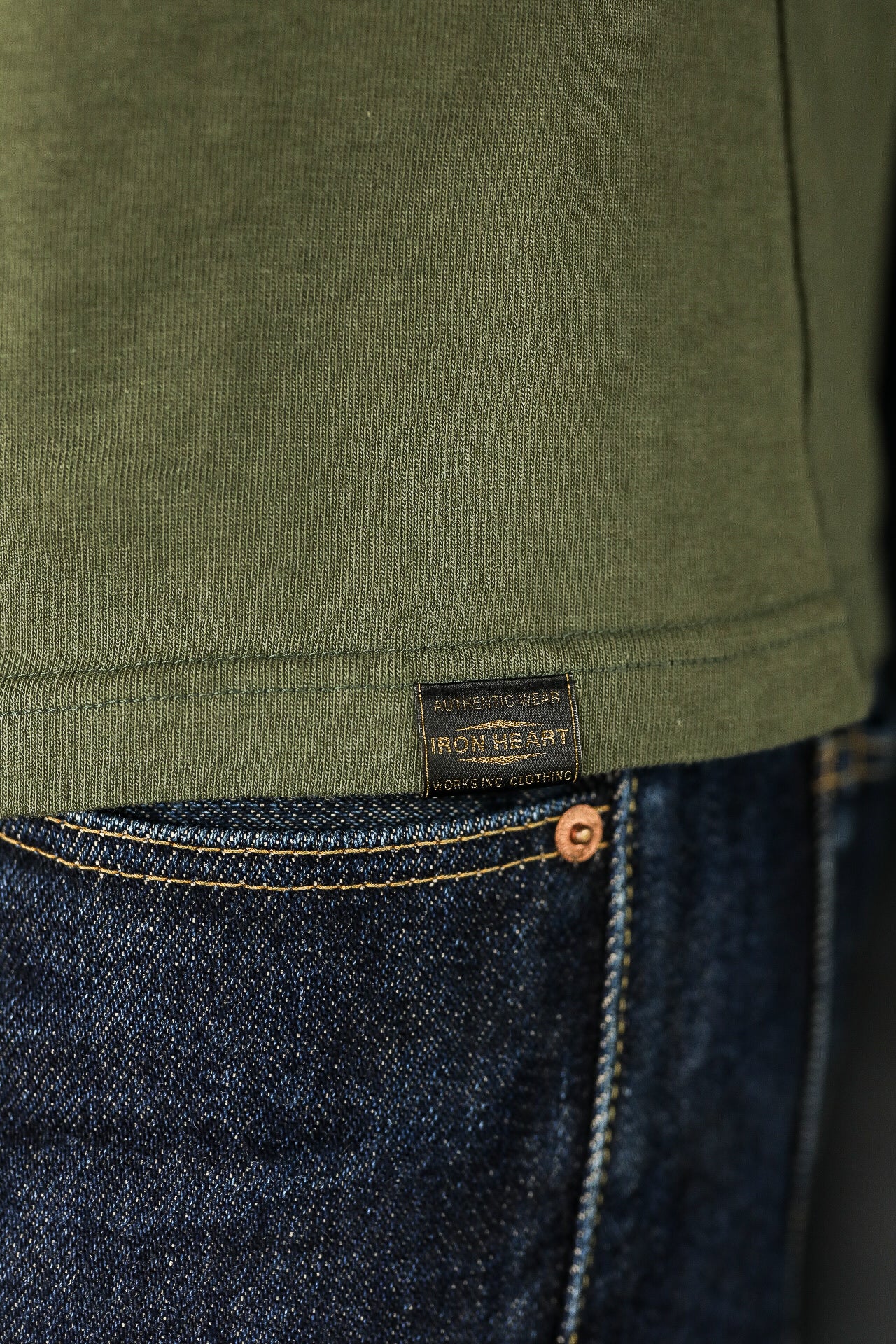 Close-up of olive green Iron Heart shirt tag above dark blue denim jeans with yellow stitching