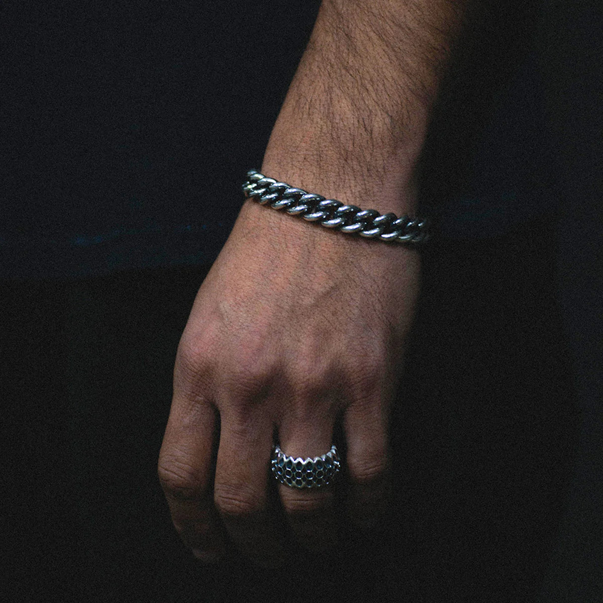 Close-up of a hand wearing a thick sterling silver curb chain bracelet and a textured silver ring against a dark background