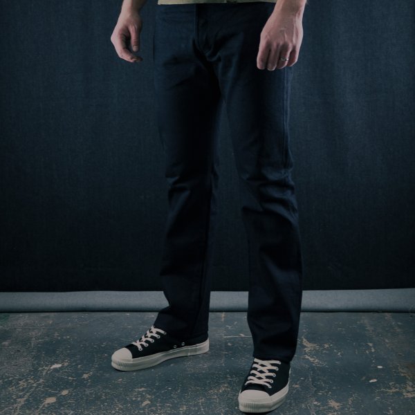 Man wearing black jeans and black lace-up sneakers standing on textured floor against dark backdrop