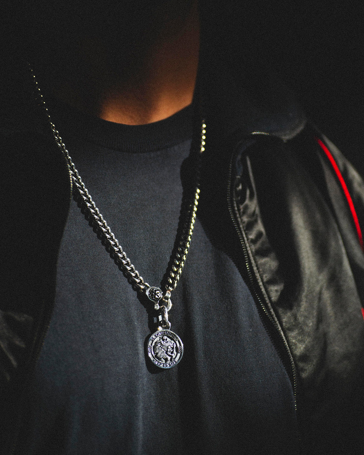 Close-up of a person wearing a thick silver chain necklace with a St. Christopher pendant over a dark shirt and jacket