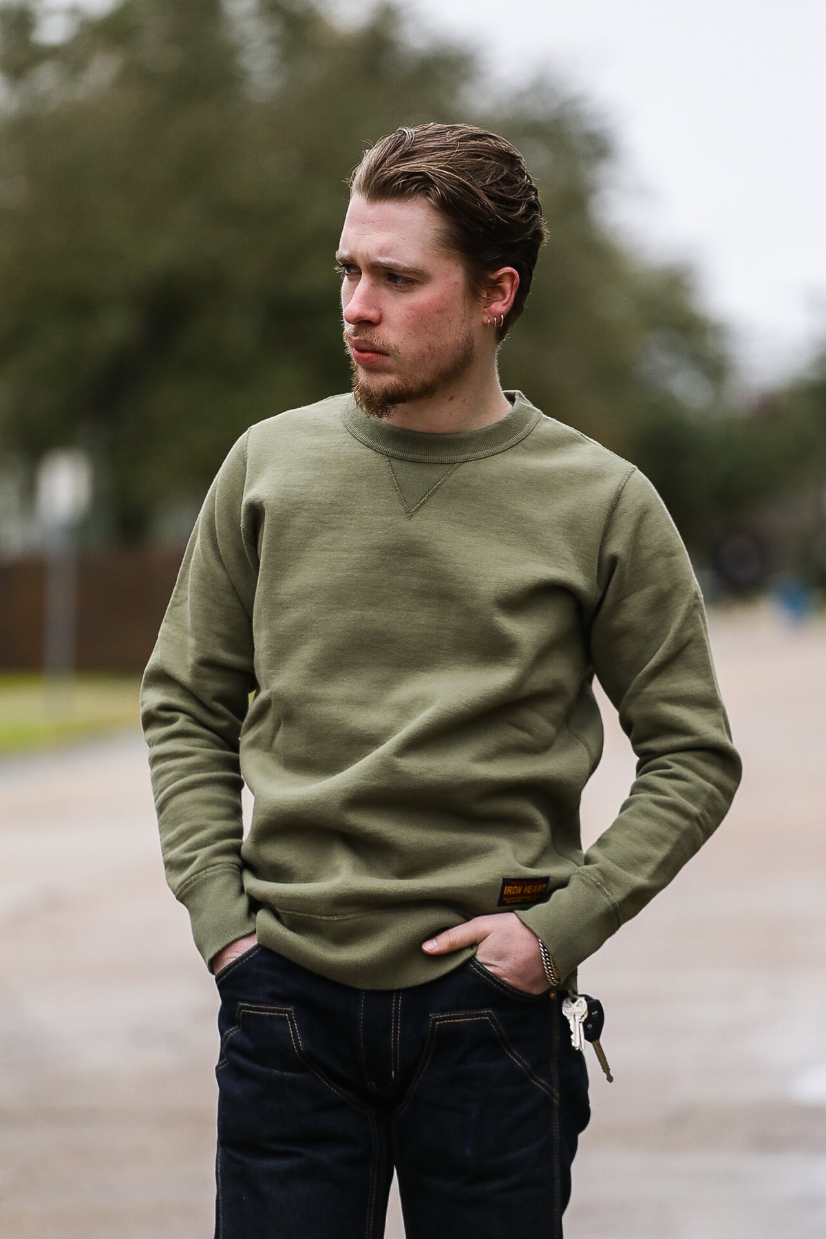 man wearing olive green crewneck sweatshirt and dark blue jeans outdoors with hands in pockets