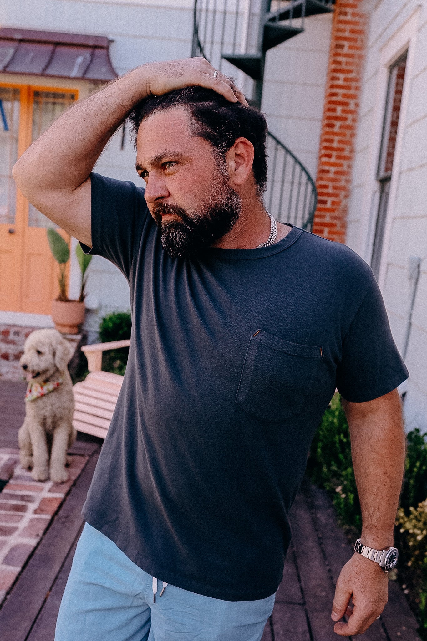 Bearded man wearing a dark pocket tee and light shorts outside near brick walkway with a dog sitting in the background