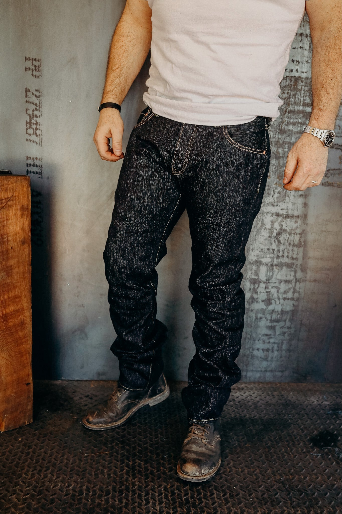 Man wearing dark raw denim jeans with rugged leather boots on industrial metal floor