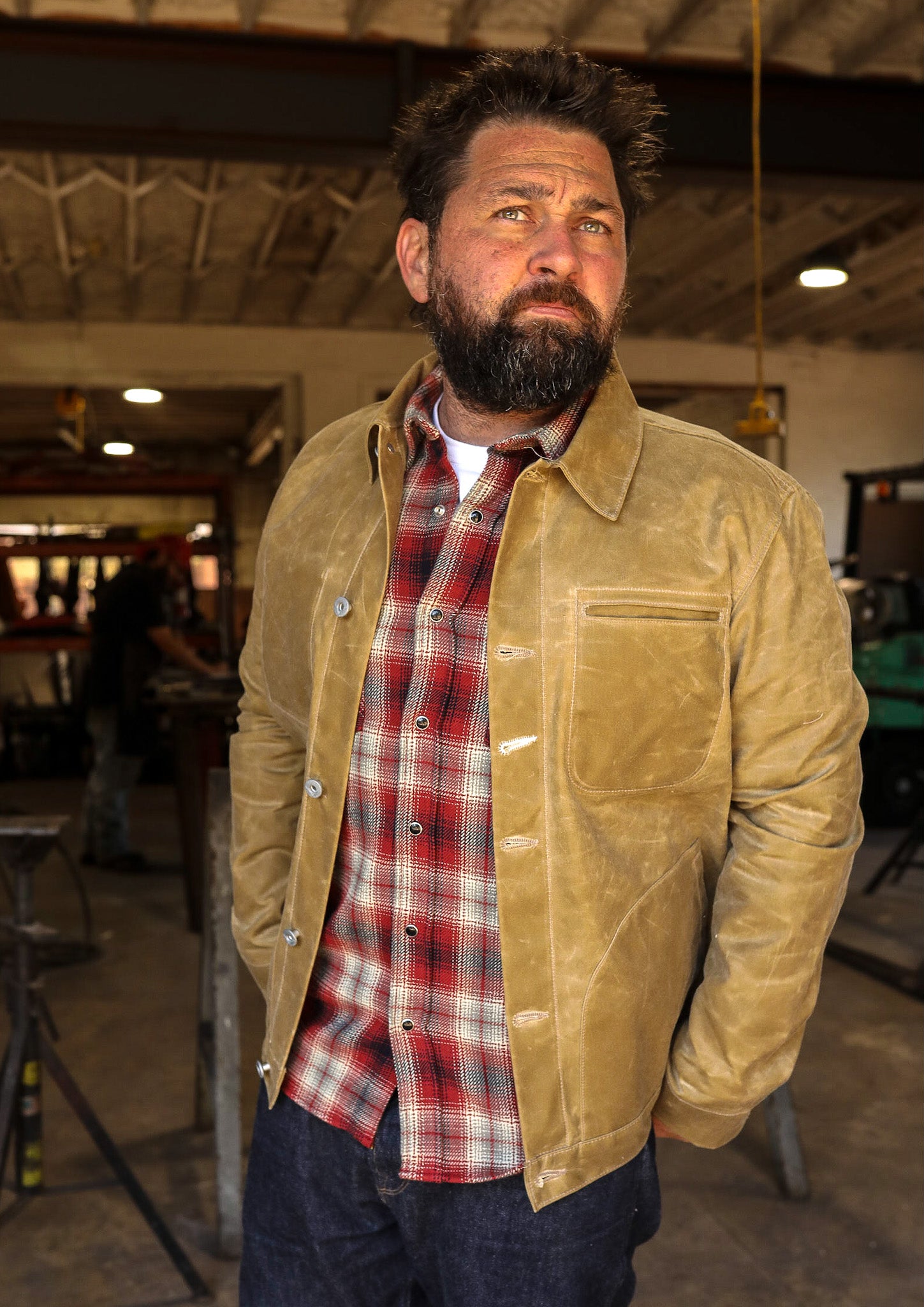 Man wearing tan leather jacket and red plaid shirt in industrial workshop setting
