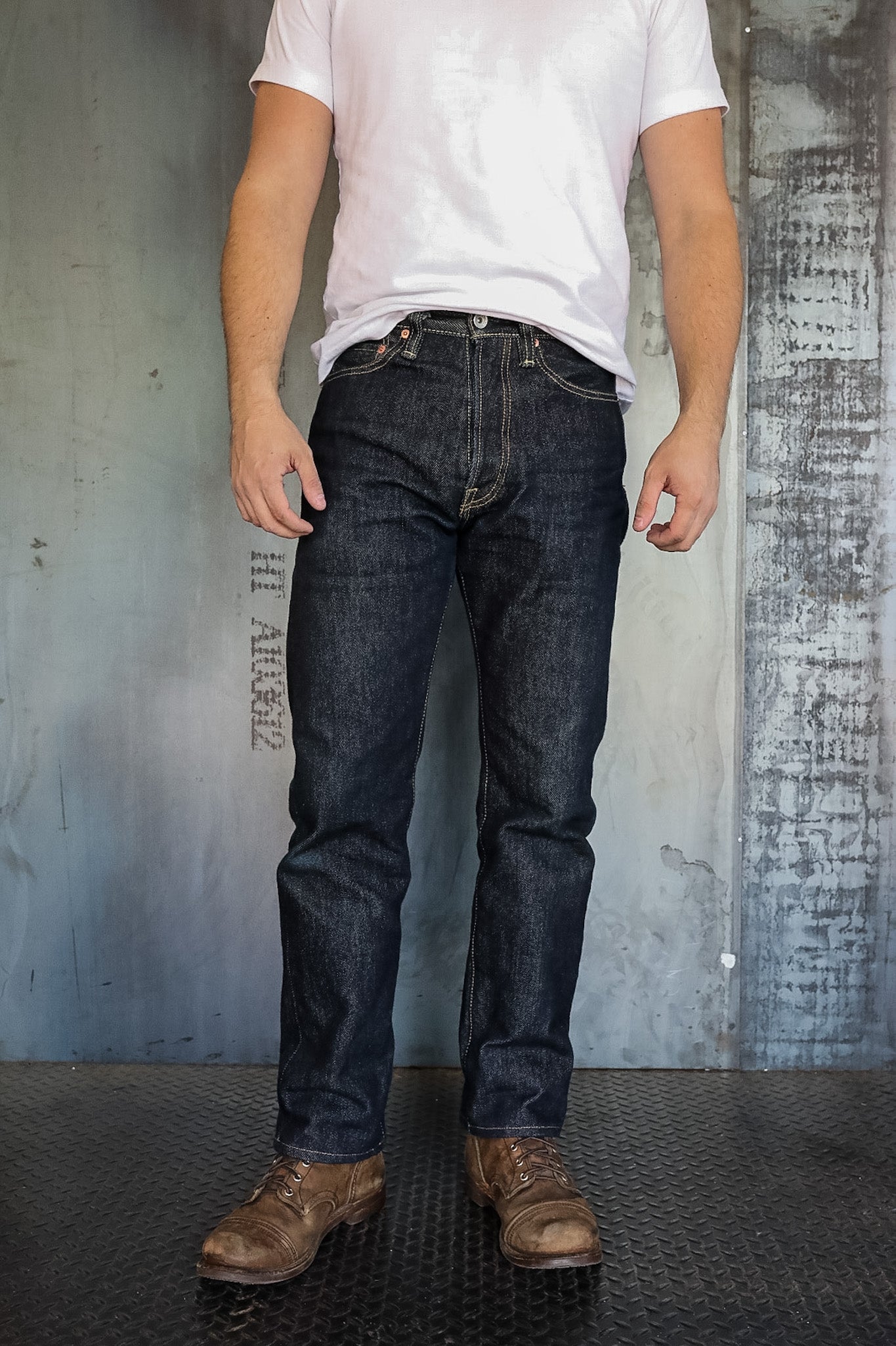 Man wearing dark raw denim jeans and brown leather boots standing on textured floor against industrial wall