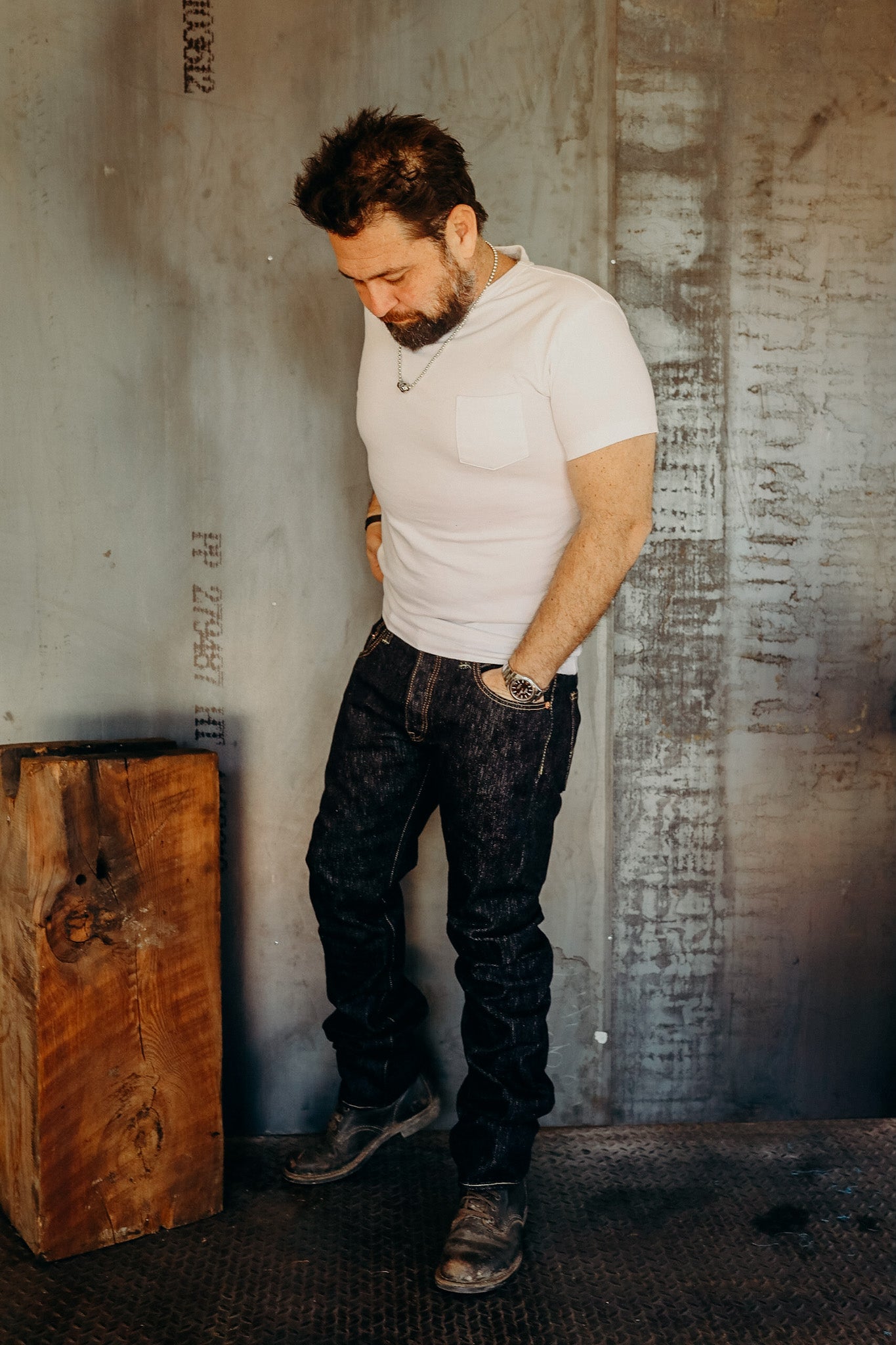 Man wearing white t-shirt, dark jeans, and rugged boots standing in industrial setting with wooden block