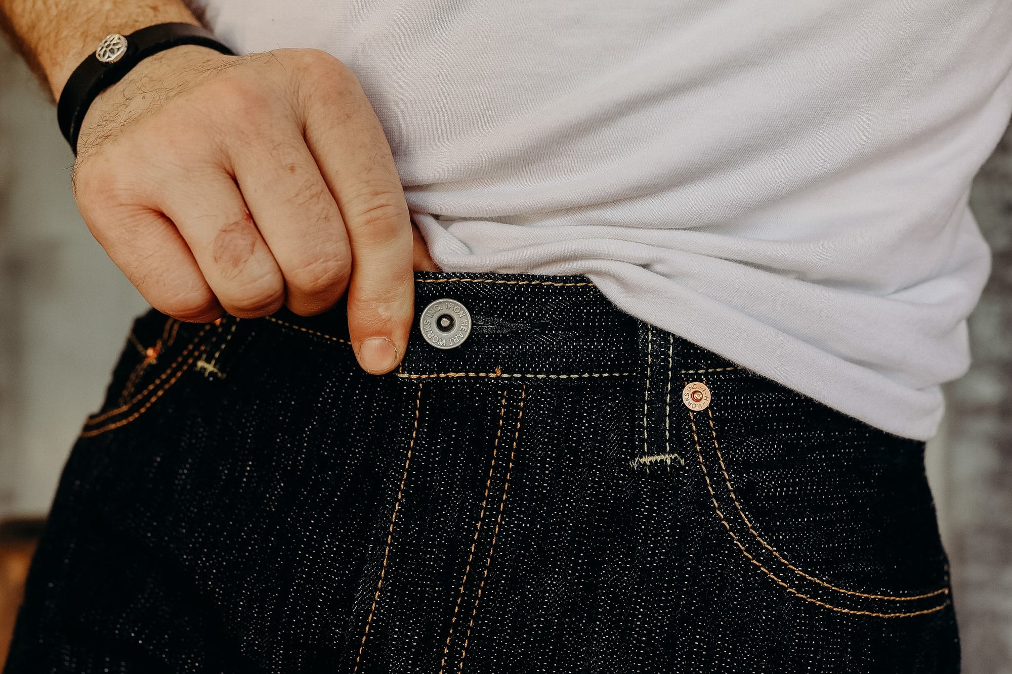 Close-up of a person fastening raw denim jeans with branded copper rivets and button, wearing a white t-shirt and black wristband