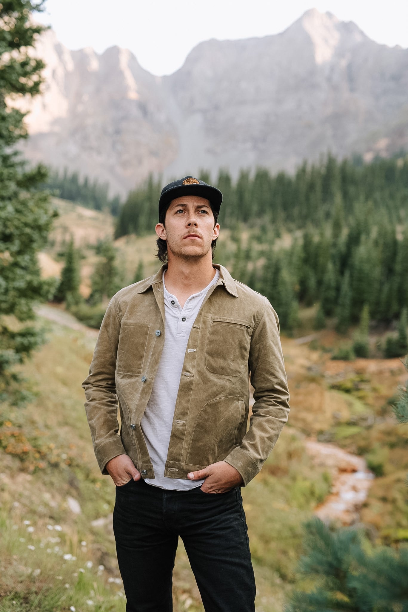 Man wearing tan canvas jacket and black cap standing in mountain forest landscape