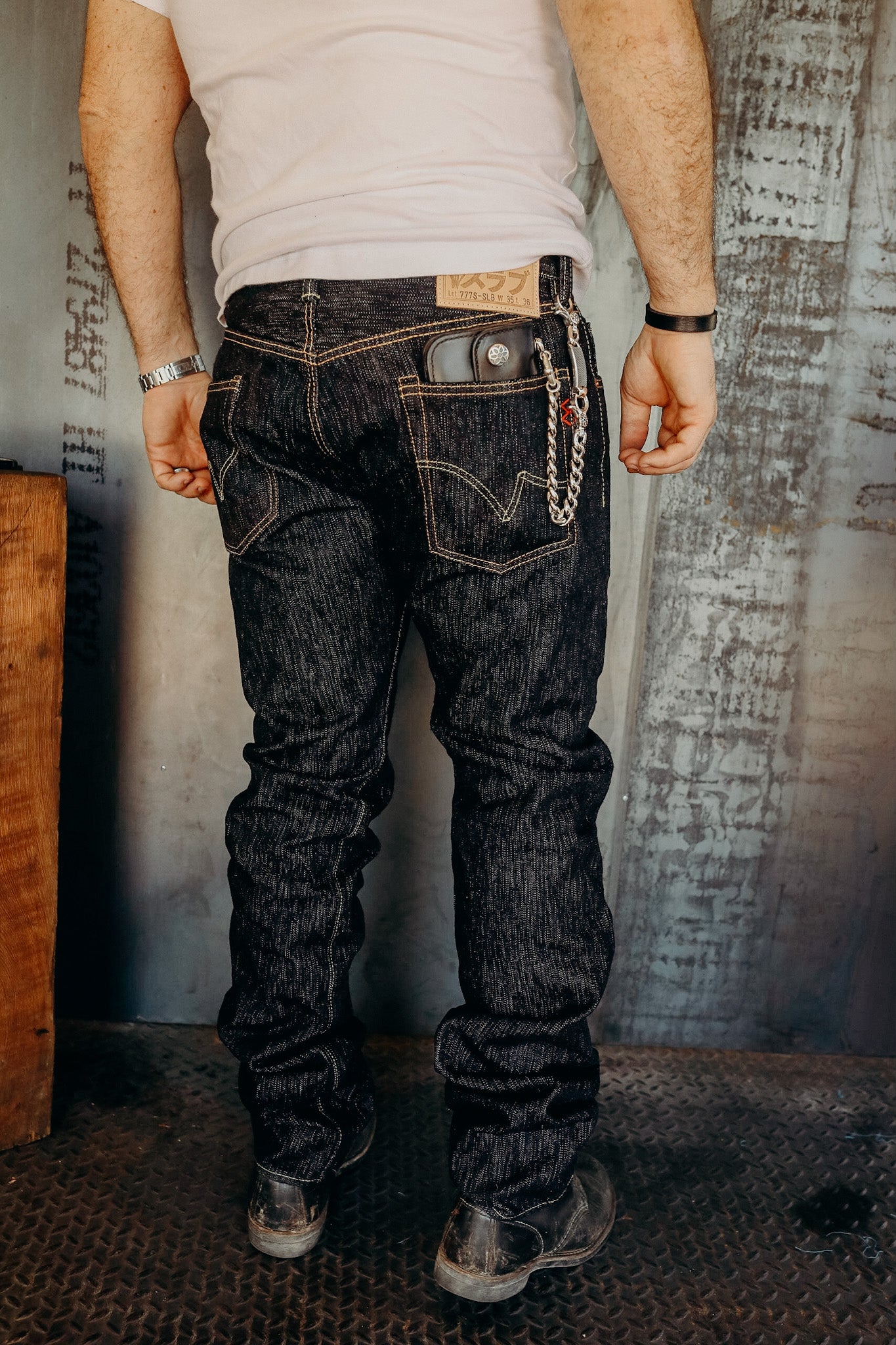 Man wearing dark raw denim jeans with wallet and chain, white tank top, and black boots against industrial wall
