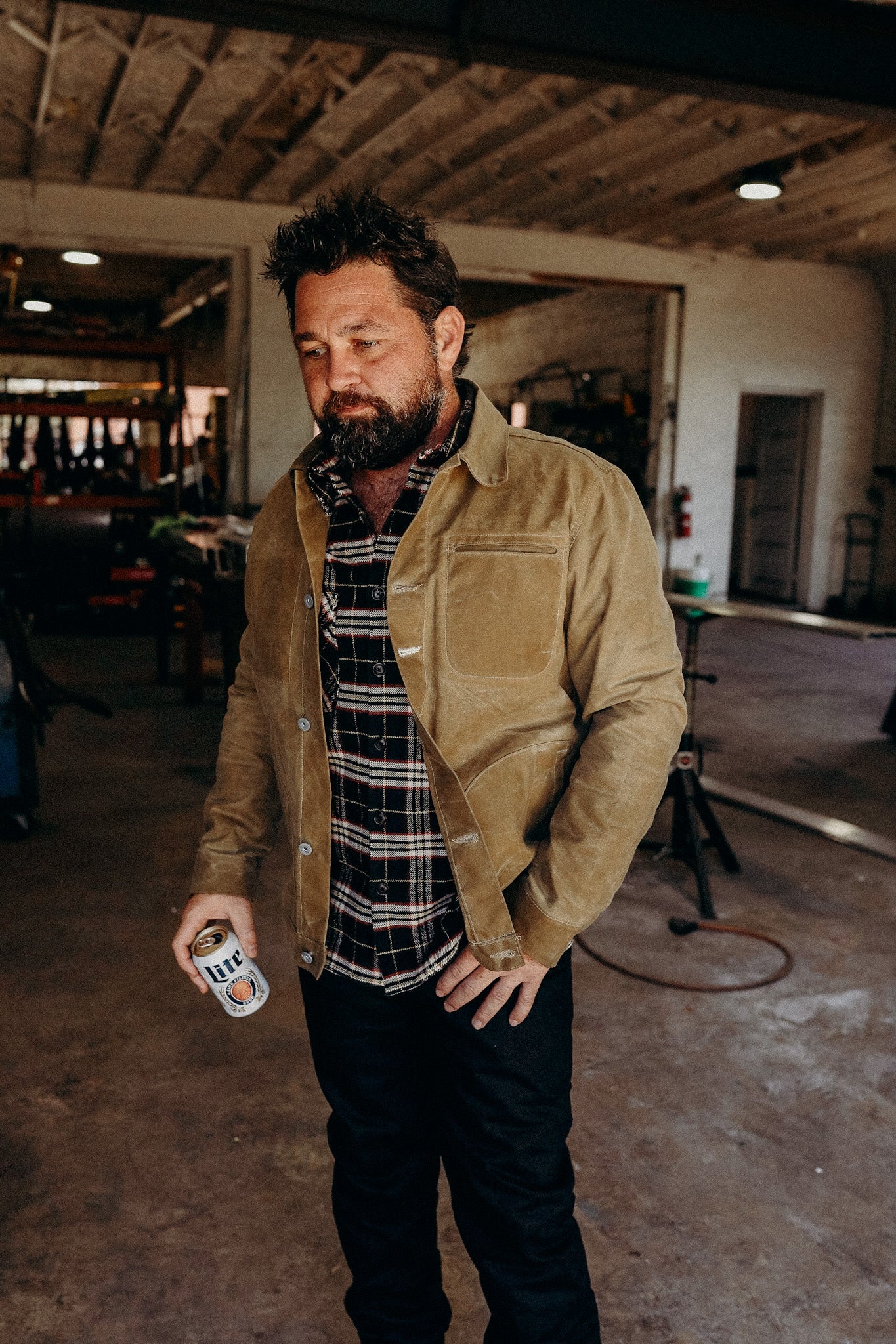 Bearded man wearing tan jacket and plaid shirt holding a can of Miller Lite in rustic workshop