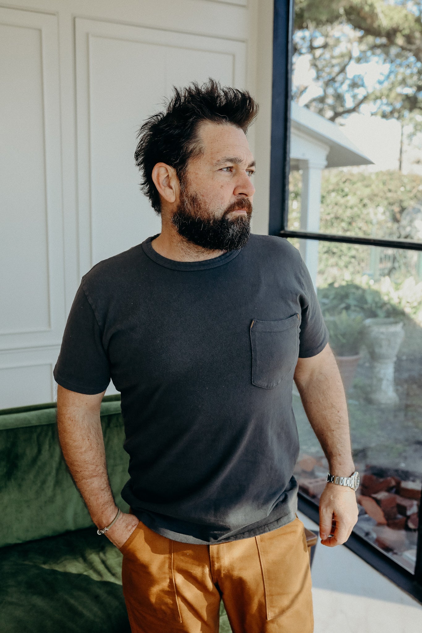 Man with beard wearing black pocket t-shirt and brown pants standing indoors near window and green couch