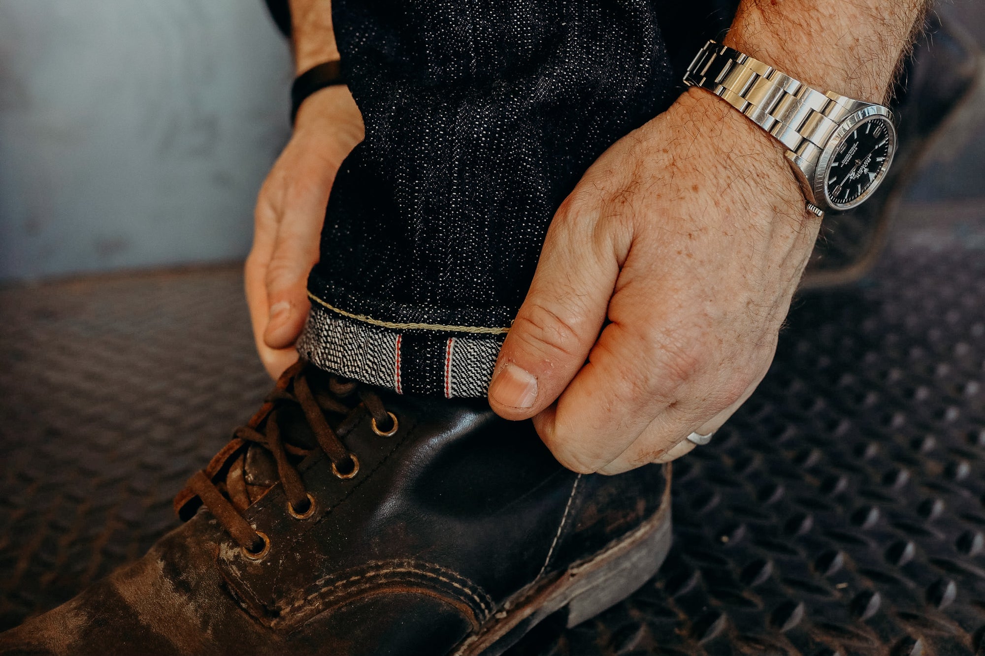 Close-up of a person wearing dark denim jeans cuffed to show selvedge edge and worn leather boots on textured metal floor