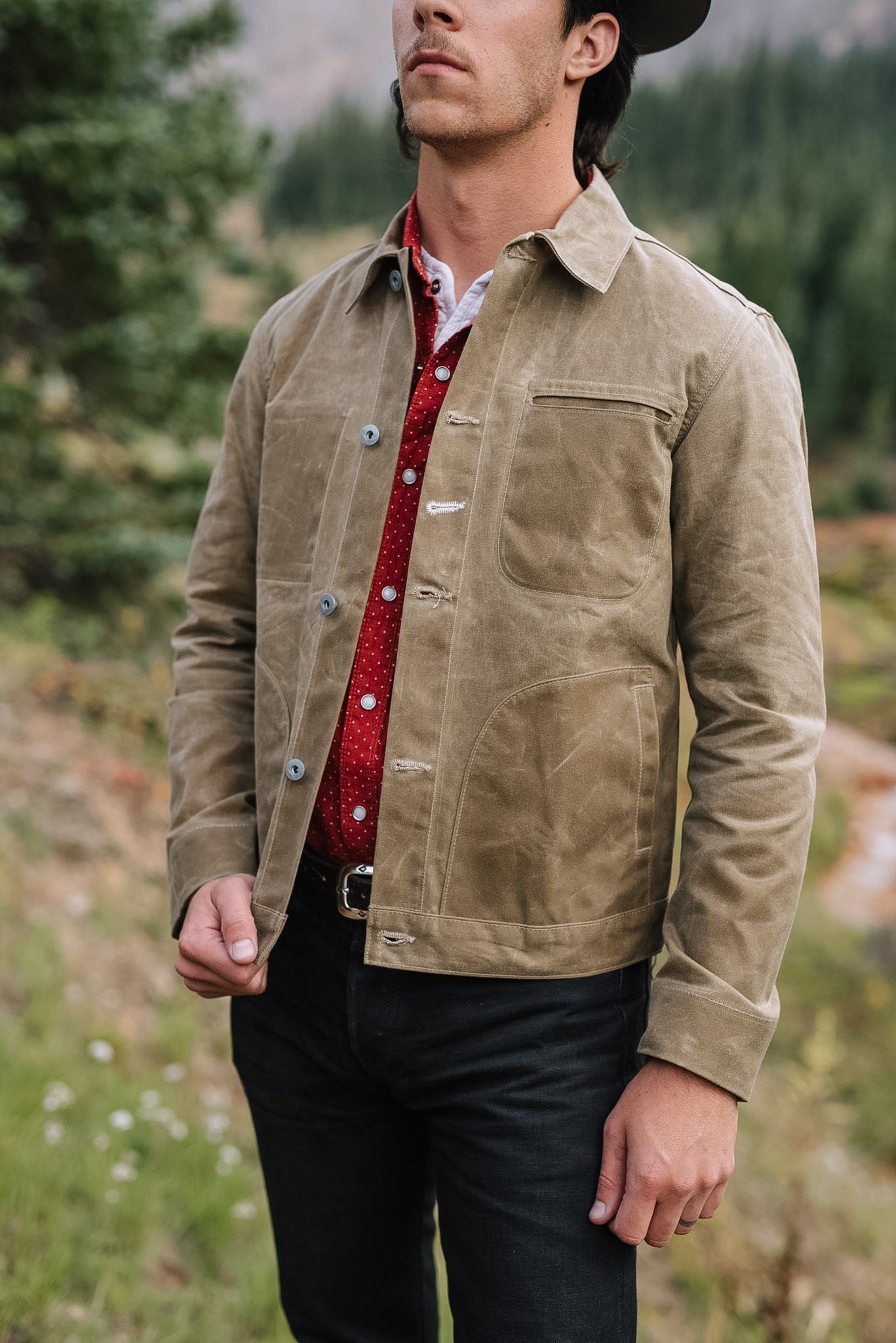 Man wearing tan canvas jacket over a red button-up shirt in a forested outdoor setting