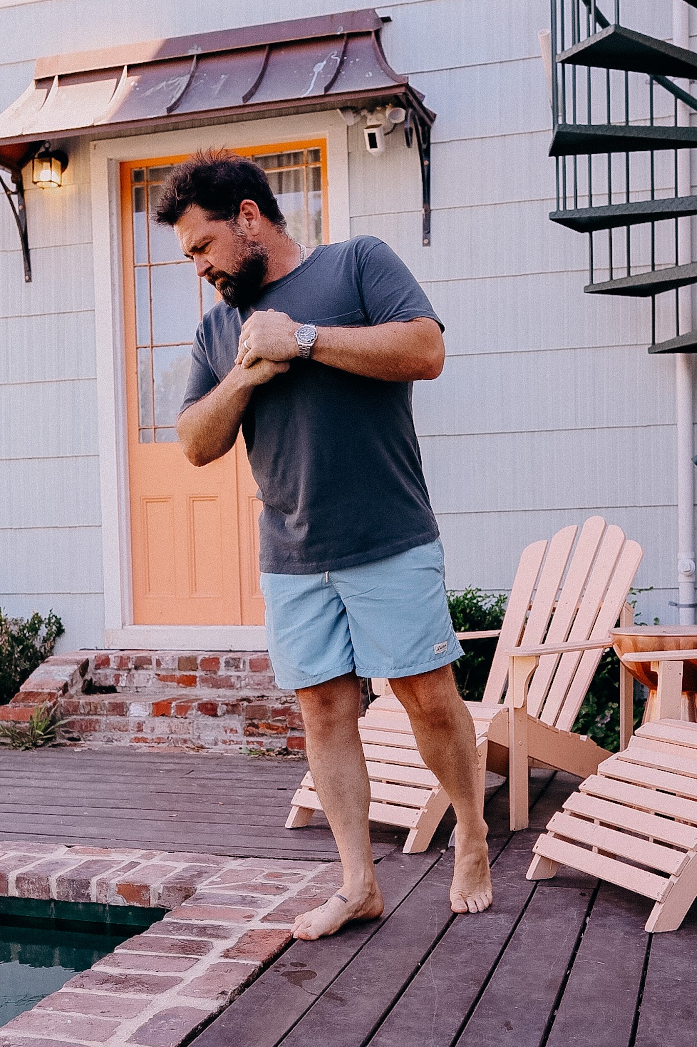 Man in gray t-shirt and light blue shorts standing barefoot on wooden deck by pool near Adirondack chairs and orange door