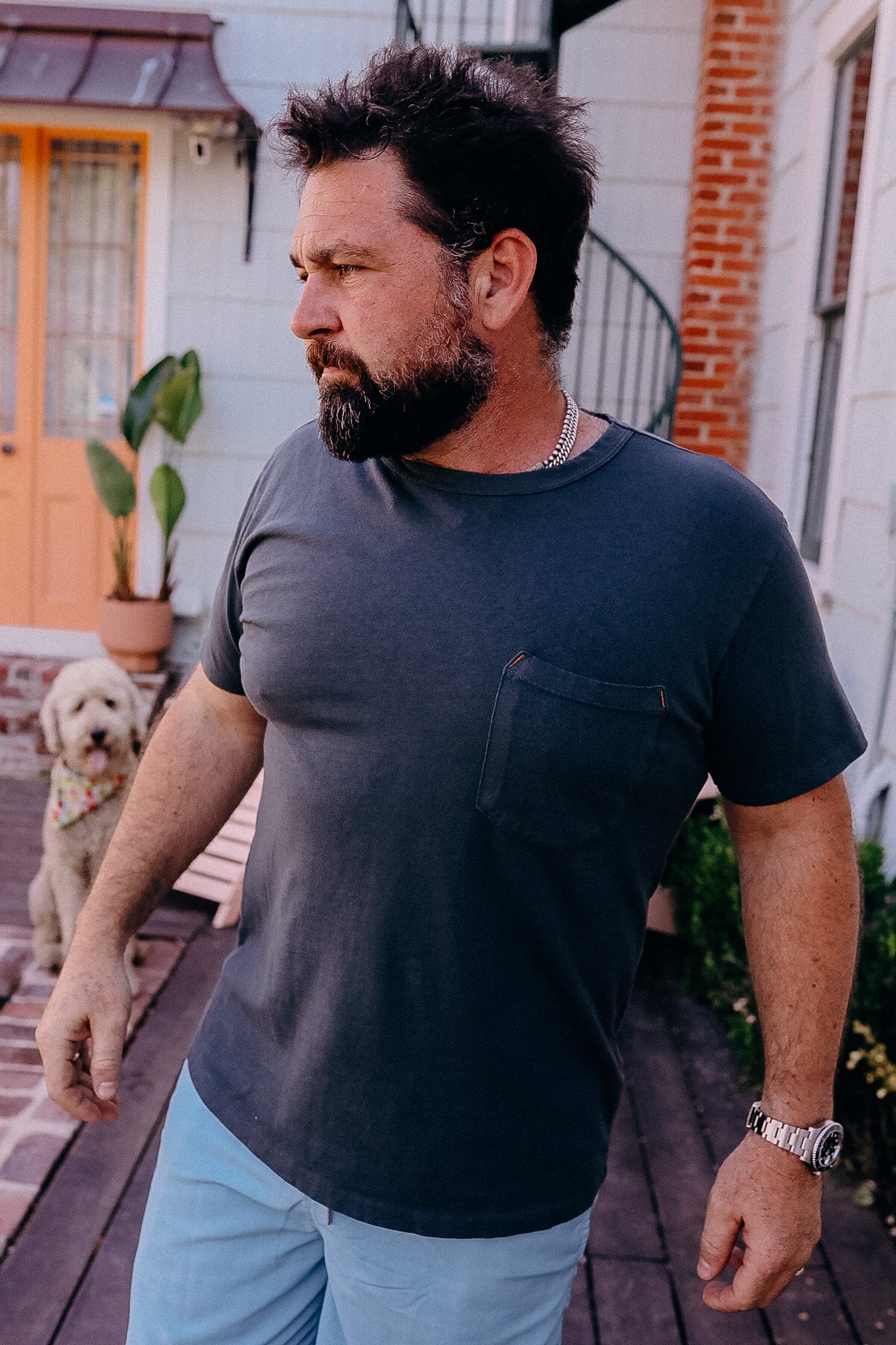 Man with beard wearing dark pocket t-shirt and light blue pants outdoors near house with dog in background